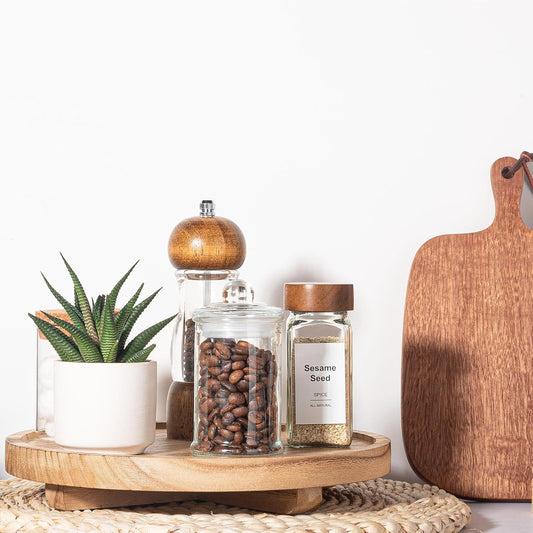 Wooden tray with a plant, pepper grinder, jar of coffee beans, and jar of sesame seeds on a white background.