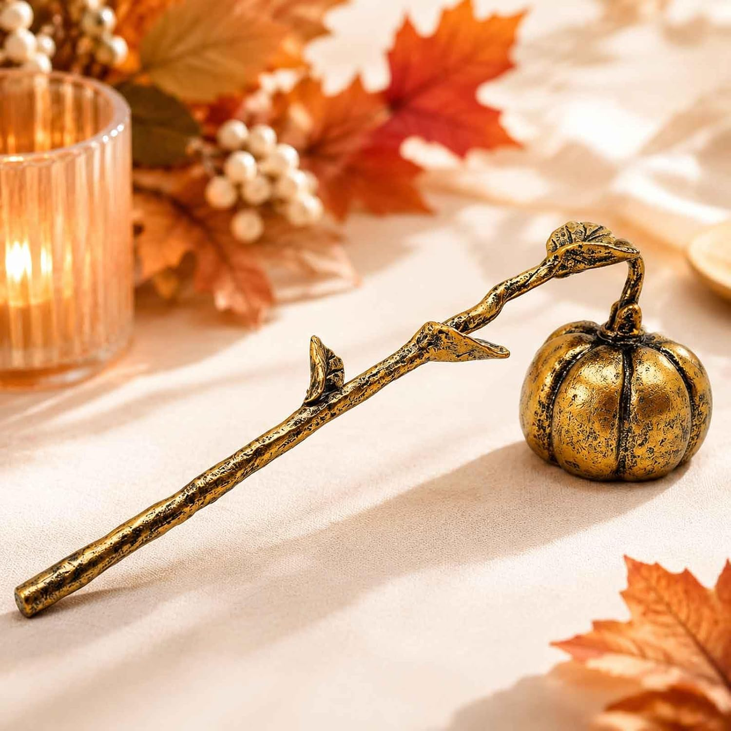 A decorative metallic branch-shaped object with a small pumpkin at the end, styled on a beige fabric surface with autumn leaves, a candle, and berries in the background.