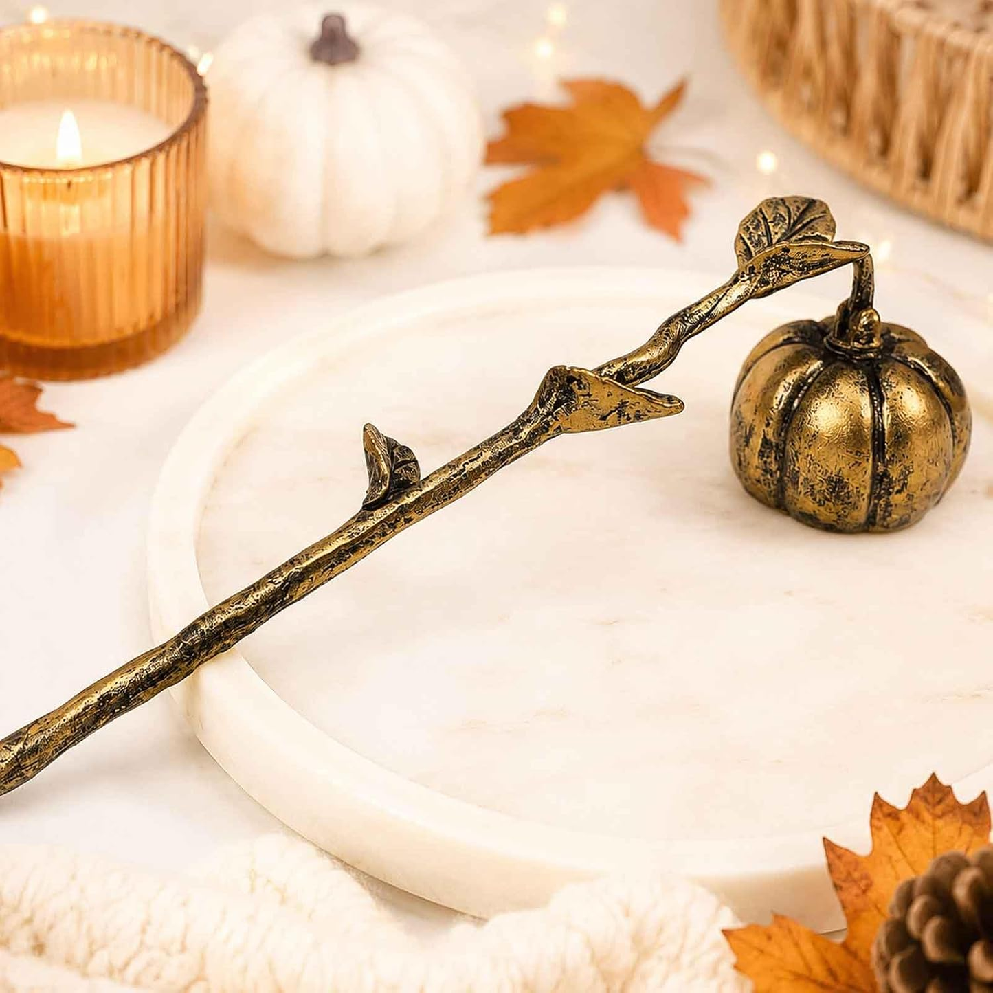 A decorative metal candle holder shaped like a branch with a small pumpkin at the end, styled on a white marble tray with a lit amber candle, white pumpkins, autumn leaves, and a knit blanket in the background.