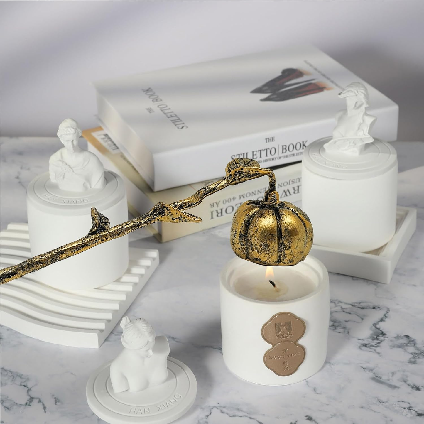 A styled product arrangement on a marble surface featuring white ceramic candle holders with sculptural white figurines, a white candle with a gold heart-shaped wax seal, a gold-toned decorative branch, and stacked books in the background.