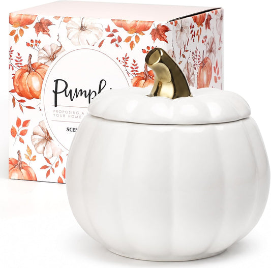 A white ceramic pumpkin-shaped scented candle jar with a lid and a metallic gold stem, shown in front of its illustrated retail box featuring pumpkins and autumn leaves.