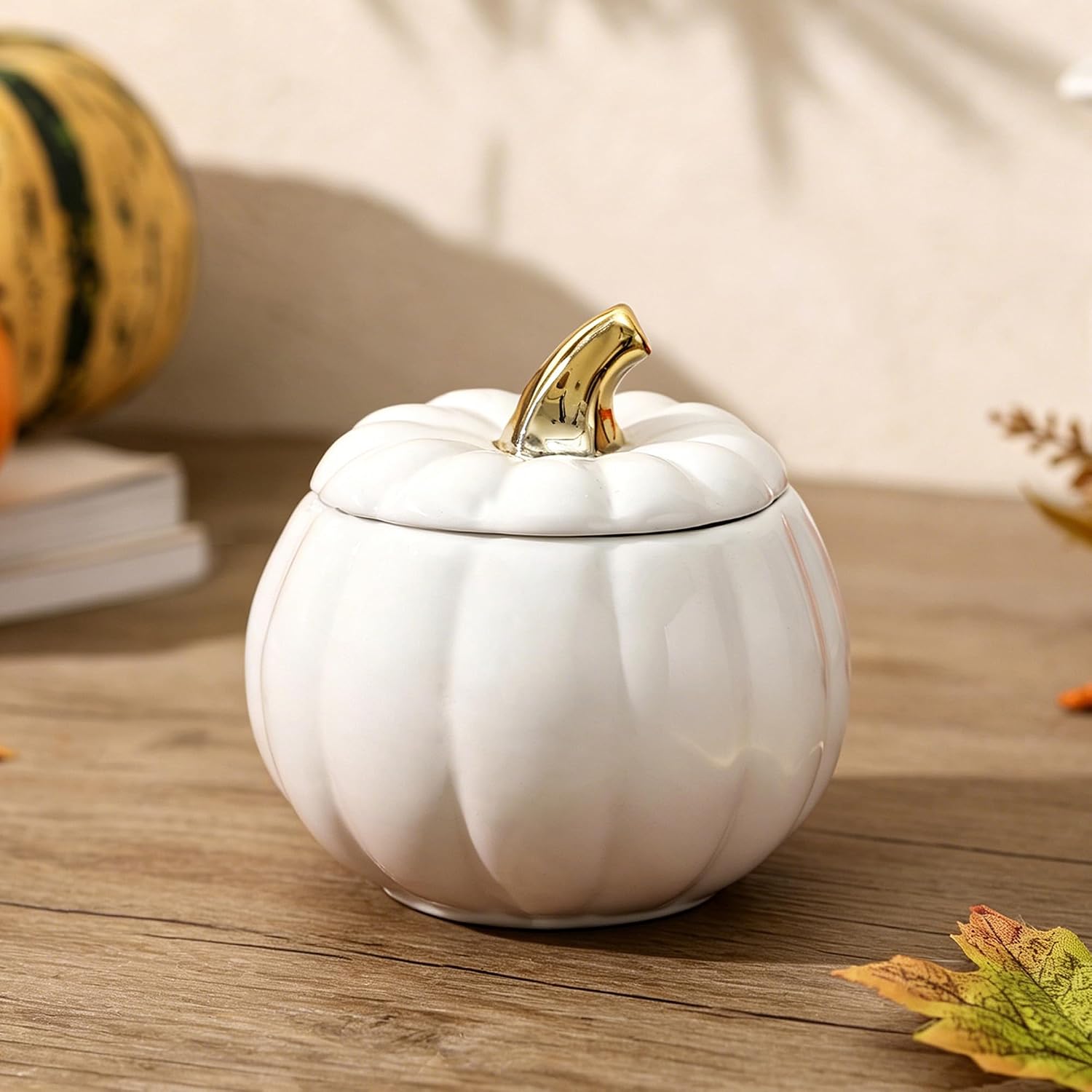 A white ceramic pumpkin-shaped lidded container with a gold-toned stem handle, placed on a wooden surface with autumn-themed props in the background.