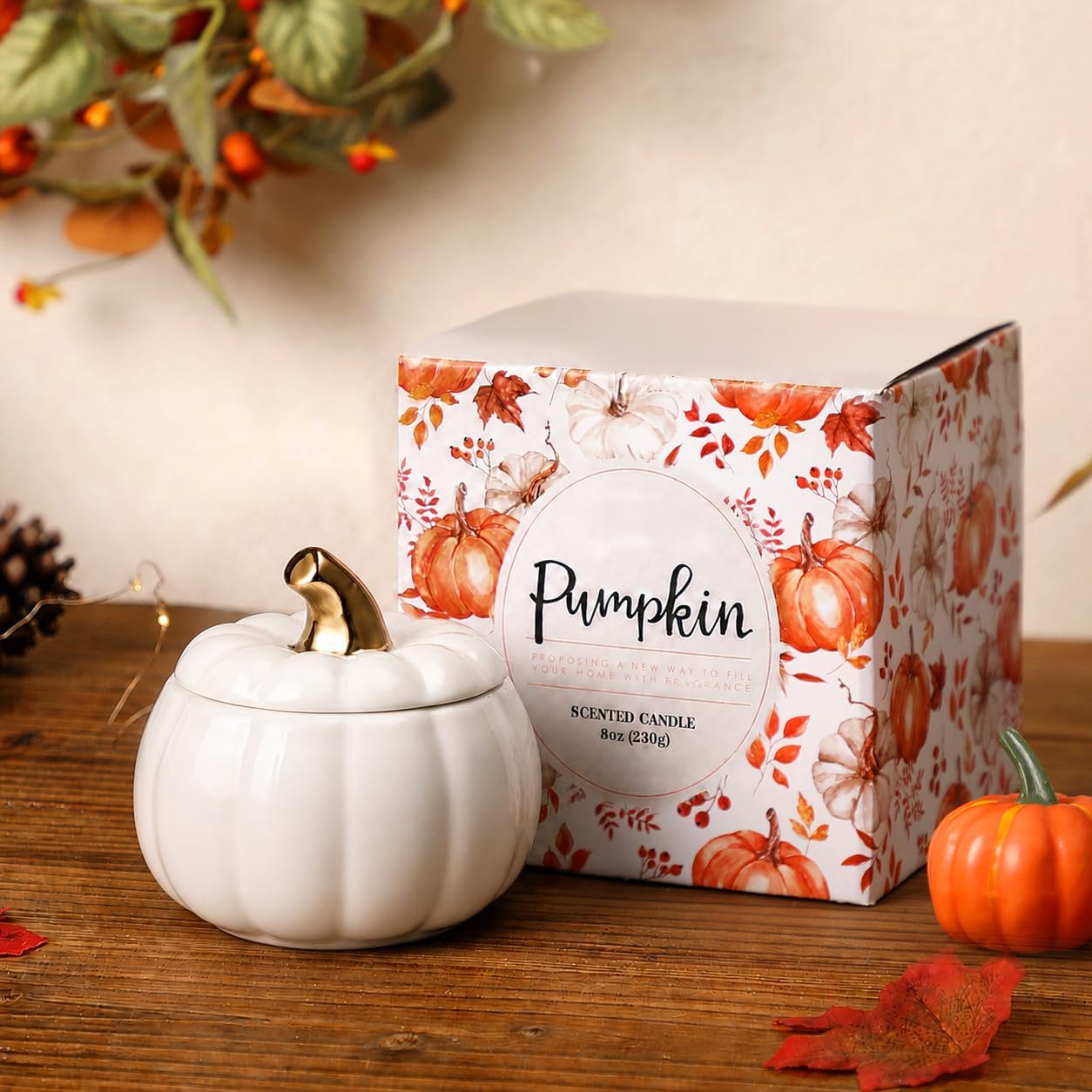 A white pumpkin-shaped scented candle jar with a gold-toned stem sits beside its printed retail box on a wooden surface. The box is white with orange and red autumn leaf and pumpkin illustrations and reads 'Pumpkin' and 'Scented Candle'.
