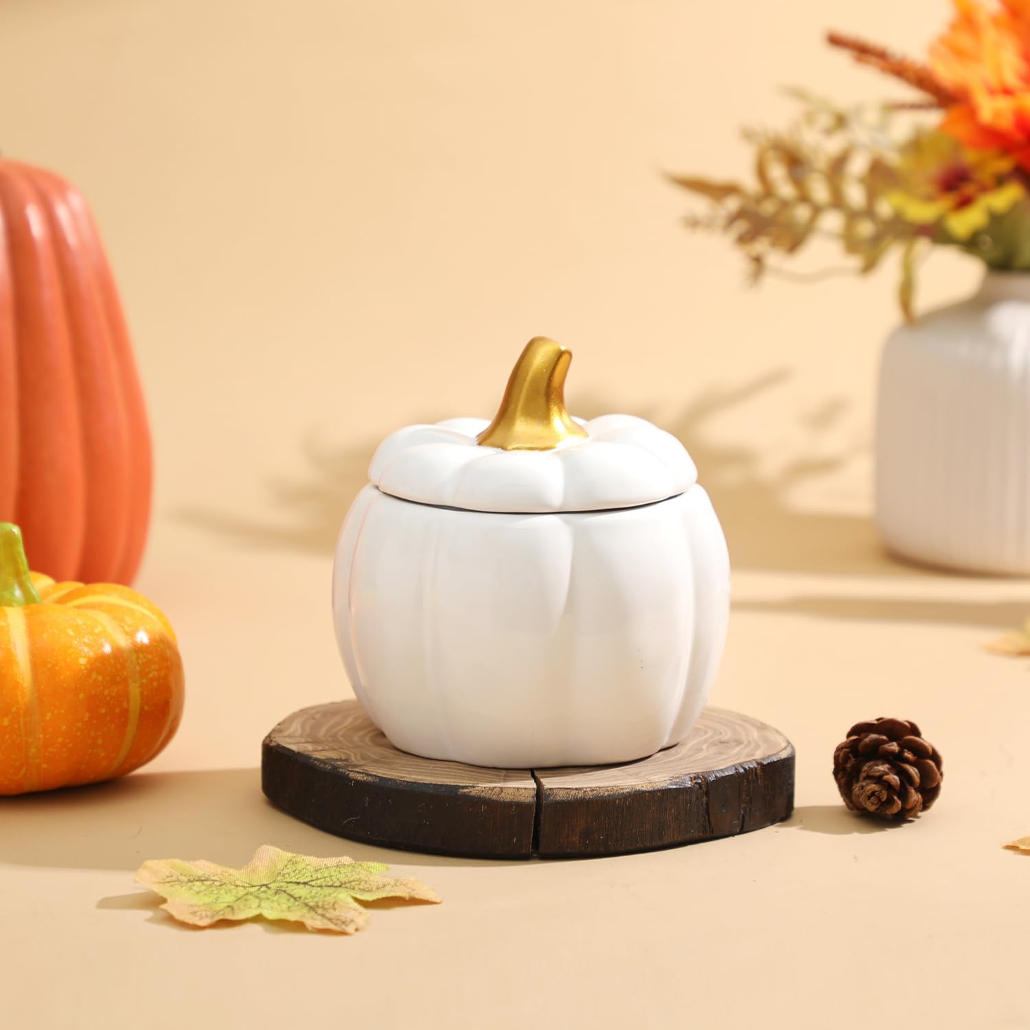 White pumpkin-shaped container with a gold lid on a wooden base, surrounded by autumn decorations.