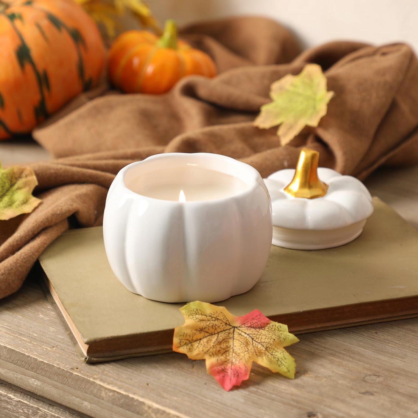 White pumpkin-shaped candle with a gold top on a wooden surface with pumpkins and leaves.