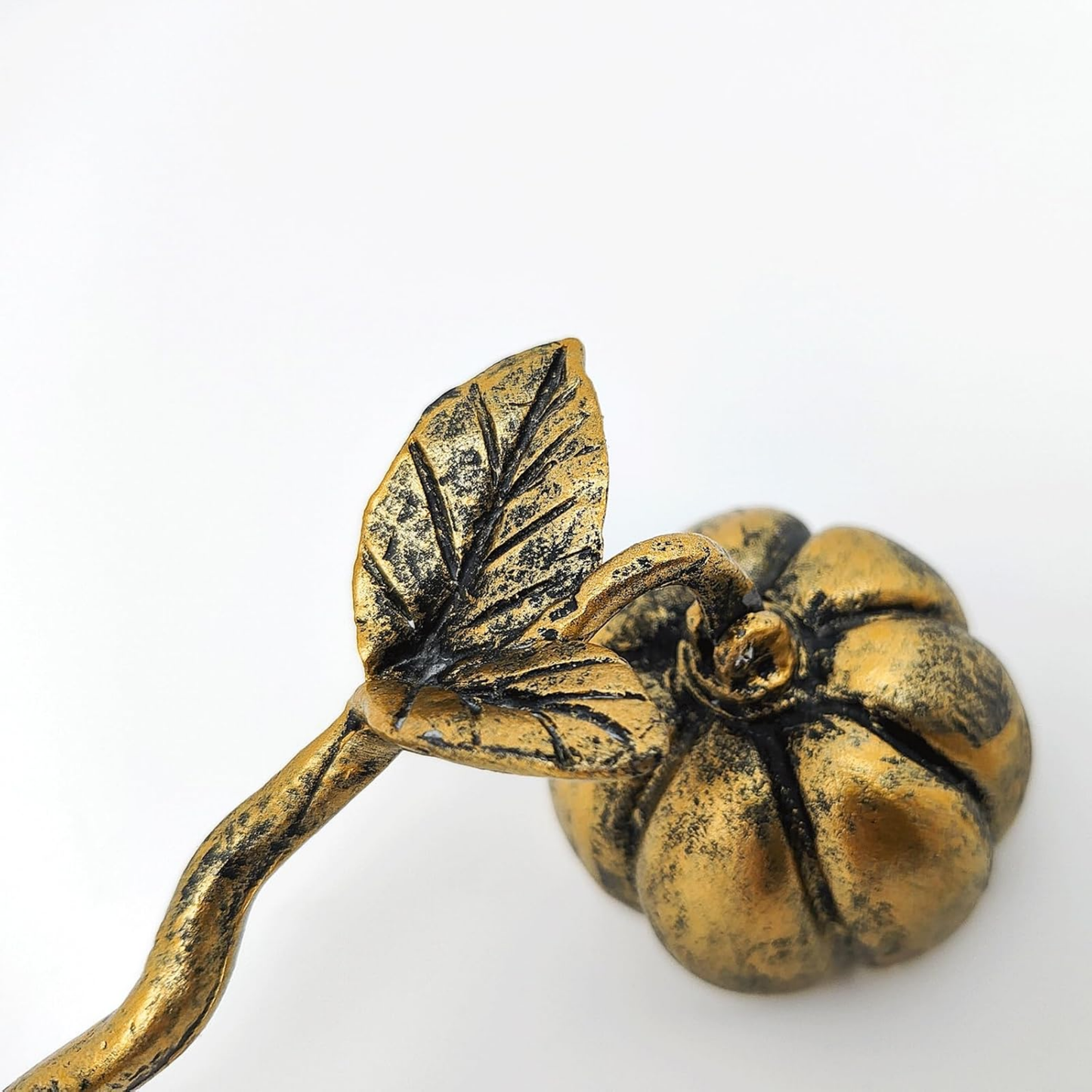 A close-up of a metallic gold decorative object with a leaf and stem leading to a pumpkin-shaped form, photographed on a plain white background.